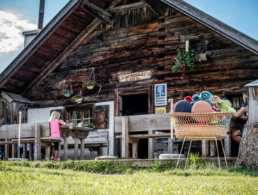 Lärchenhütte A rustic wooden cabin hosts Alpine summer events, with people at tables and towels stacked in a basket nearby.