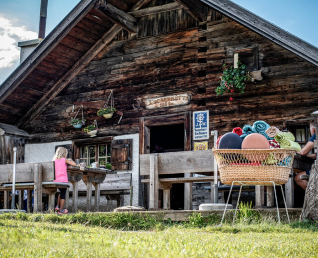 A rustic wooden cabin hosts Alpine summer events, with people at tables and towels stacked in a basket nearby.