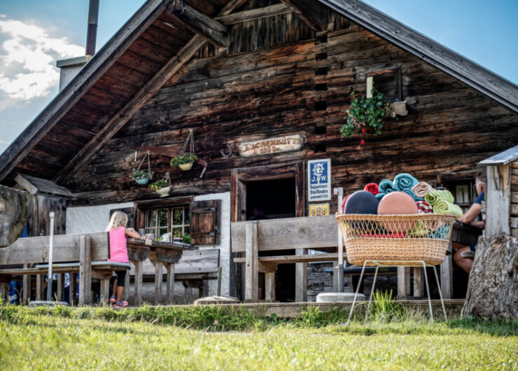 A rustic wooden cabin hosts Alpine summer events, with people at tables and towels stacked in a basket nearby.