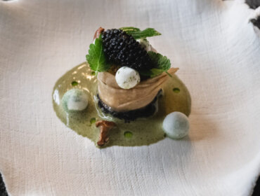 A gourmet dish with caviar, greens, and foam on a textured white plate against a dark background.