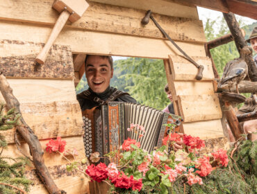 Smiling person playing accordion in a wooden hut decorated with flowers, with another person in the background.