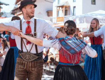 People in traditional Austrian clothing dance outdoors at a lively festival with onlookers in the background.