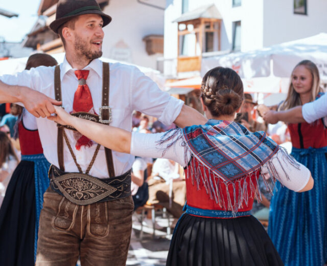 People in traditional Austrian clothing dance outdoors at a lively festival with onlookers in the background.