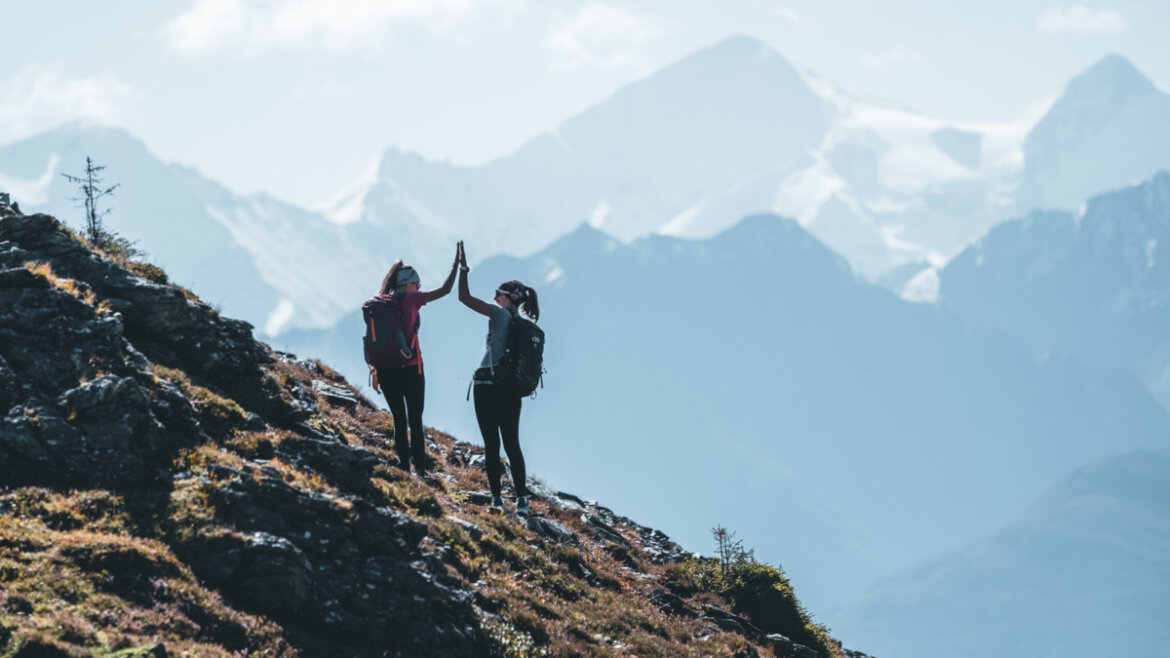 Two young women hiking on a slope with snow-covered mountains in the background.
