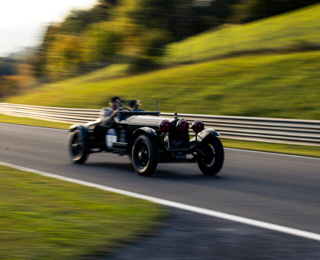 A vintage black car speeds down a racetrack with blurred background, emphasising its motion.