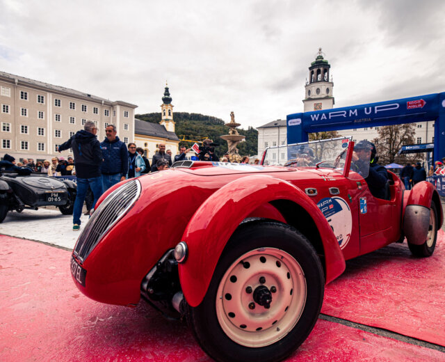 A vintage red racing car on display at an outdoor event with a crowd and historic buildings in the background.