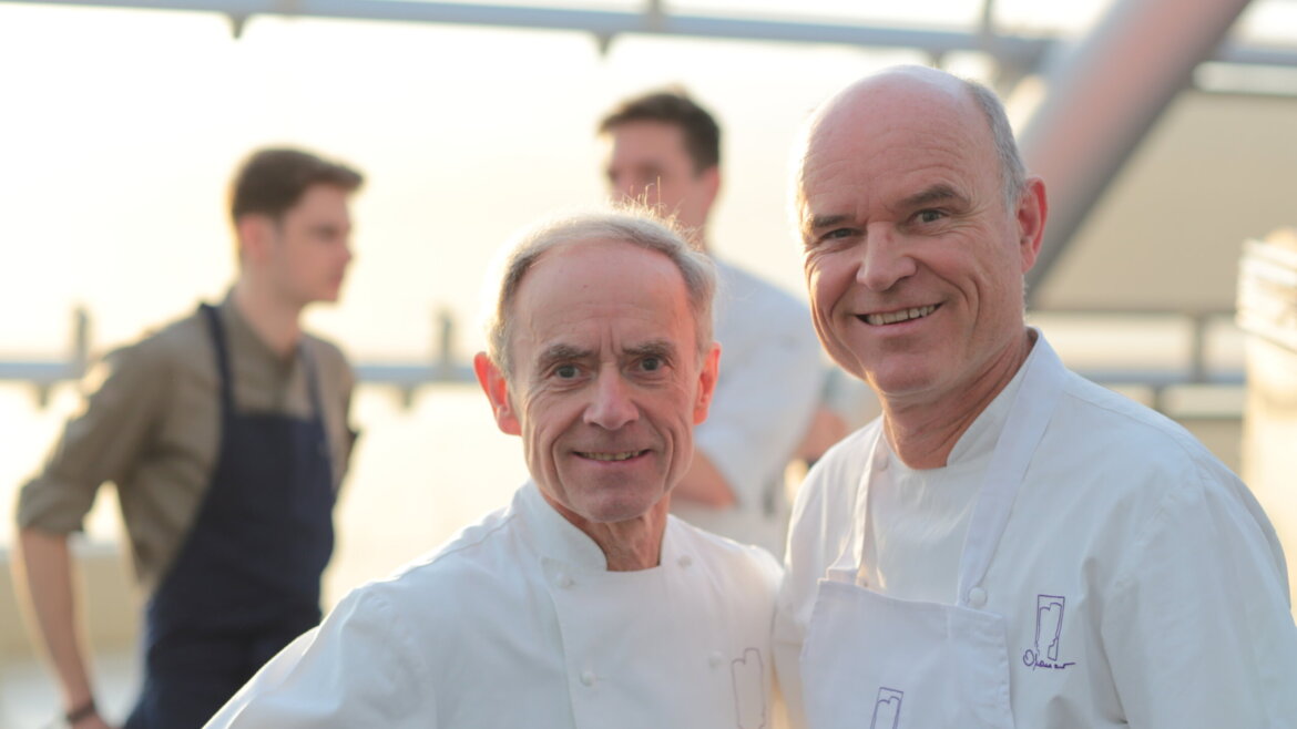 Two smiling chefs in white uniforms stand together, with two blurred people in the background. (Enlarged view)
