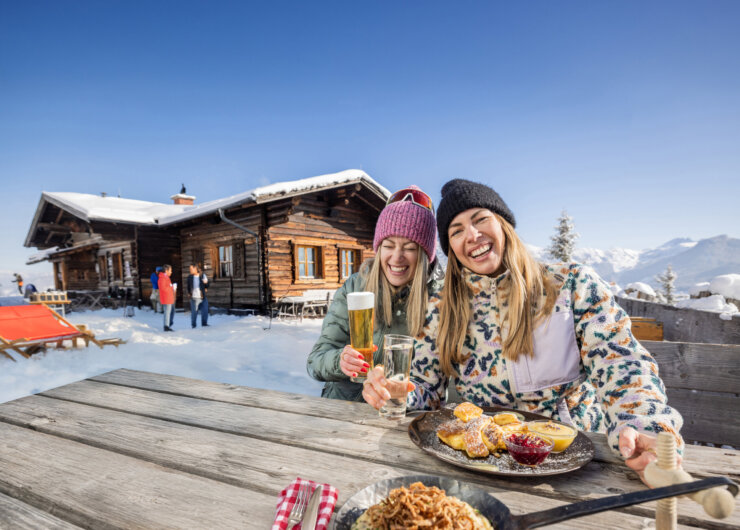 Two women sitting in front of a wooden hut in winter enjoying a meal