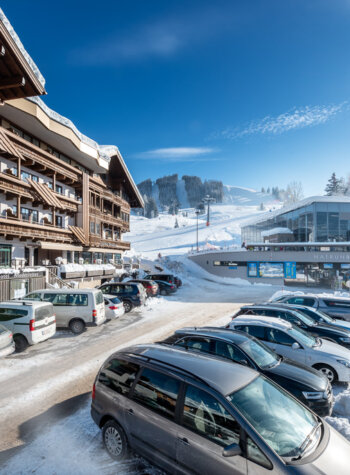 Snowy ski resort with parked cars, chalet-style buildings, and a ski lift station under a clear blue sky.