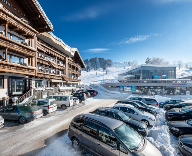Snowy ski resort with parked cars, chalet-style buildings, and a ski lift station under a clear blue sky.