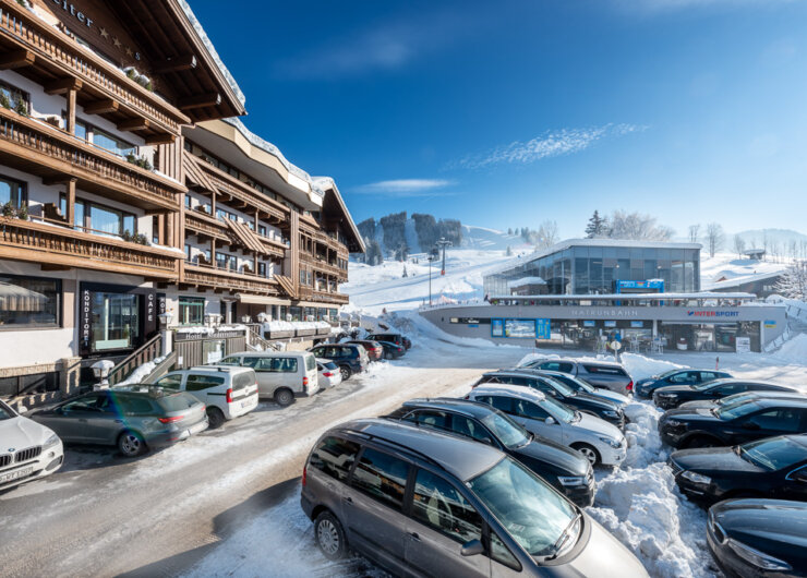 Snowy ski resort with parked cars, chalet-style buildings, and a ski lift station under a clear blue sky.