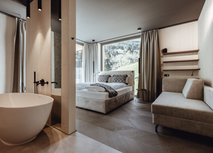 Modern bedroom with large window, freestanding bath, beige sofa, and scenic mountain view outside.