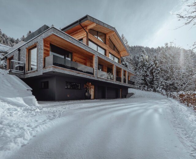 Modern chalet-style house with wood and glass, surrounded by snow and pine trees on a winter’s day.