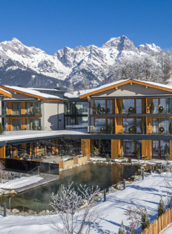 Modern alpine hotel with balconies, set by a pond, surrounded by snowy mountains and clear blue sky.