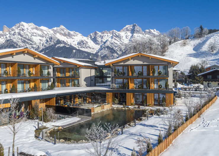 Modern alpine hotel with balconies, set by a pond, surrounded by snowy mountains and clear blue sky.
