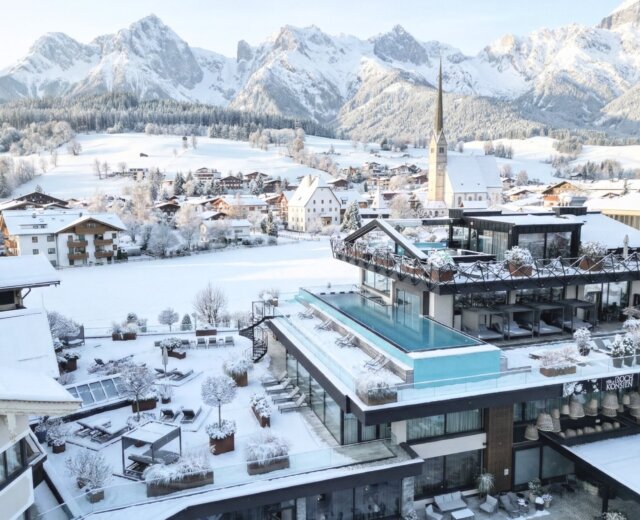 A modern hotel with a rooftop swimming pool overlooks a snowy alpine village and mountains in winter.
