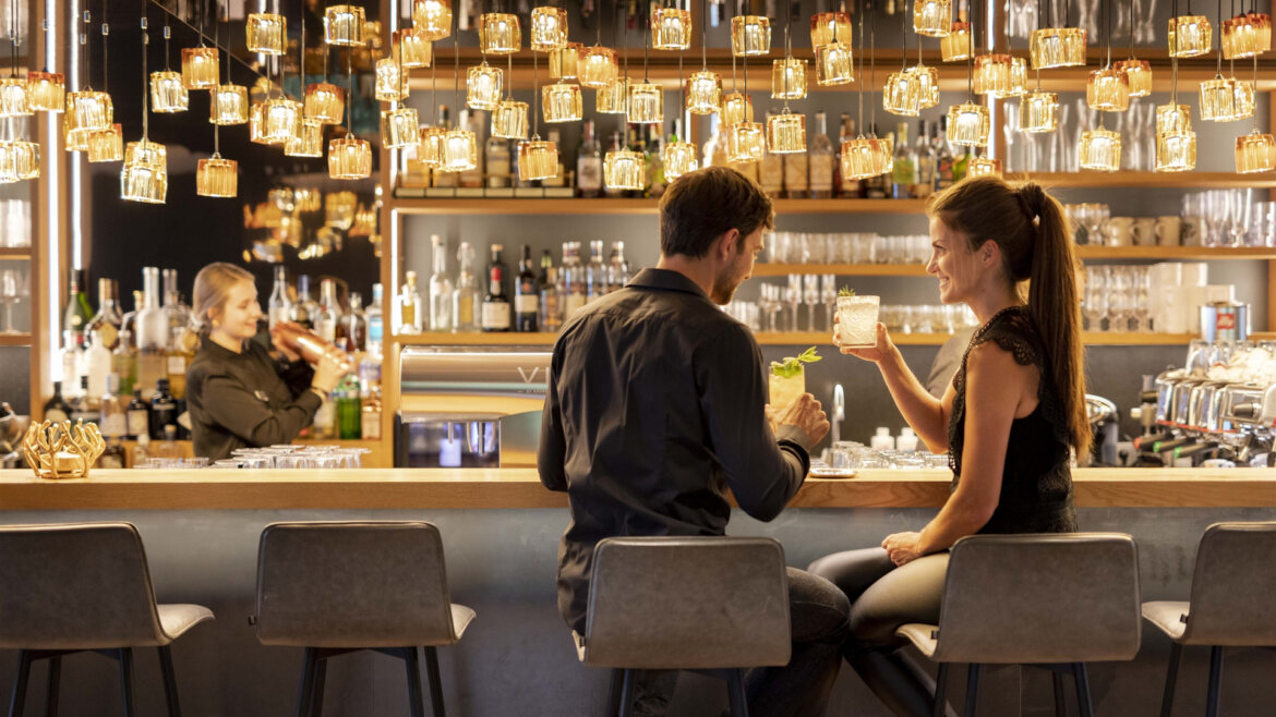 A man and woman enjoy drinks at Naturhotel Edelweiss Wagrain’s stylish, warmly lit bar with a barman behind. (Enlarged view)