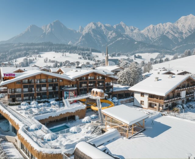 A snowy alpine resort with wooden buildings, swimming pools, and mountains in the background under a clear blue sky.