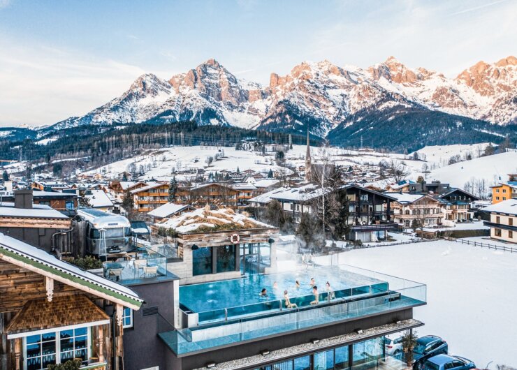 Outdoor rooftop swimming pool with people swimming, snowy village, and mountains in the background.
