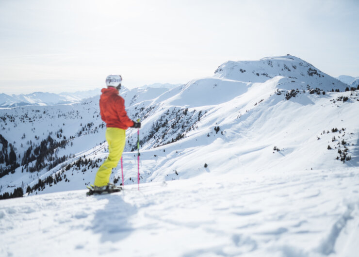 A skier in bright kit stands on a snowy mountain slope, overlooking the surrounding snow-covered peaks.