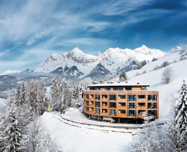 A modern wooden hotel stands amid snowy trees with towering, snow-covered mountains in the background.