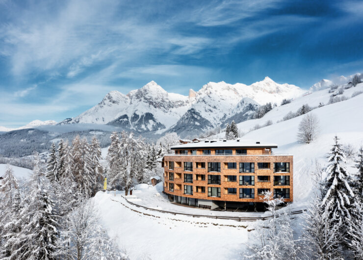 A modern wooden hotel stands amid snowy trees with towering, snow-covered mountains in the background.