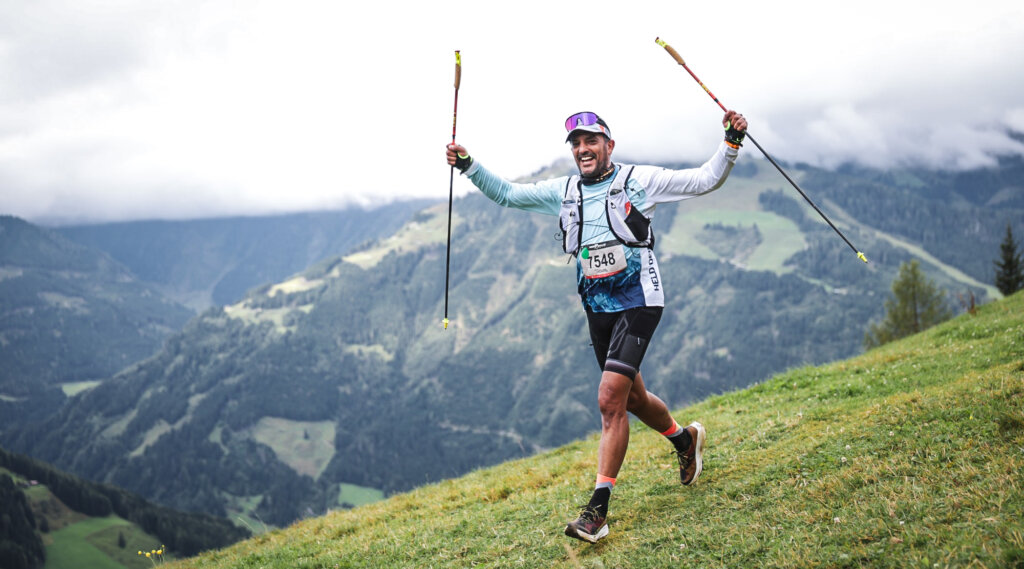Mountain Man Runner smiling and raising trekking poles on a grassy hill with mountains and clouds in the background. (Enlarged view)
