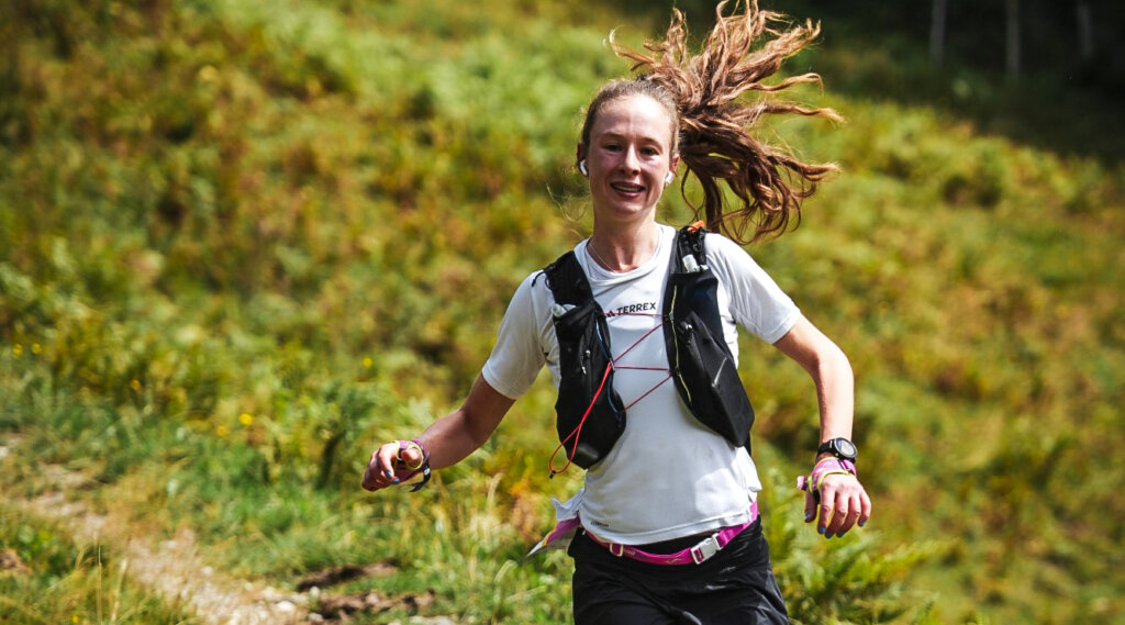 Mountainman Smiling woman fell running on a grassy path, wearing a white shirt and hydration vest. (Enlarged view)