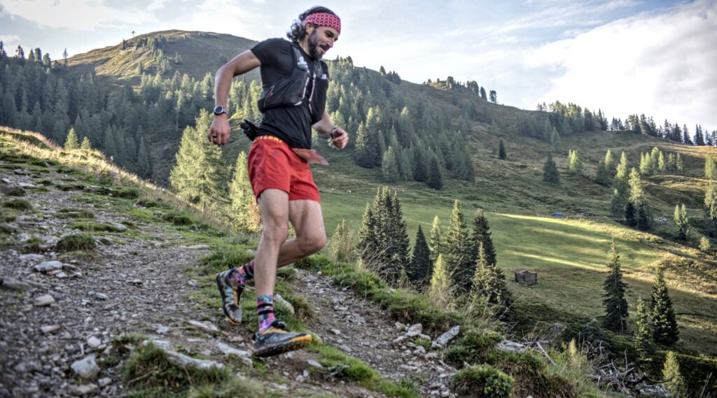 Mountain Man Trail runner in red shorts and headband descends a rocky mountain path surrounded by greenery and trees. (Enlarged view)