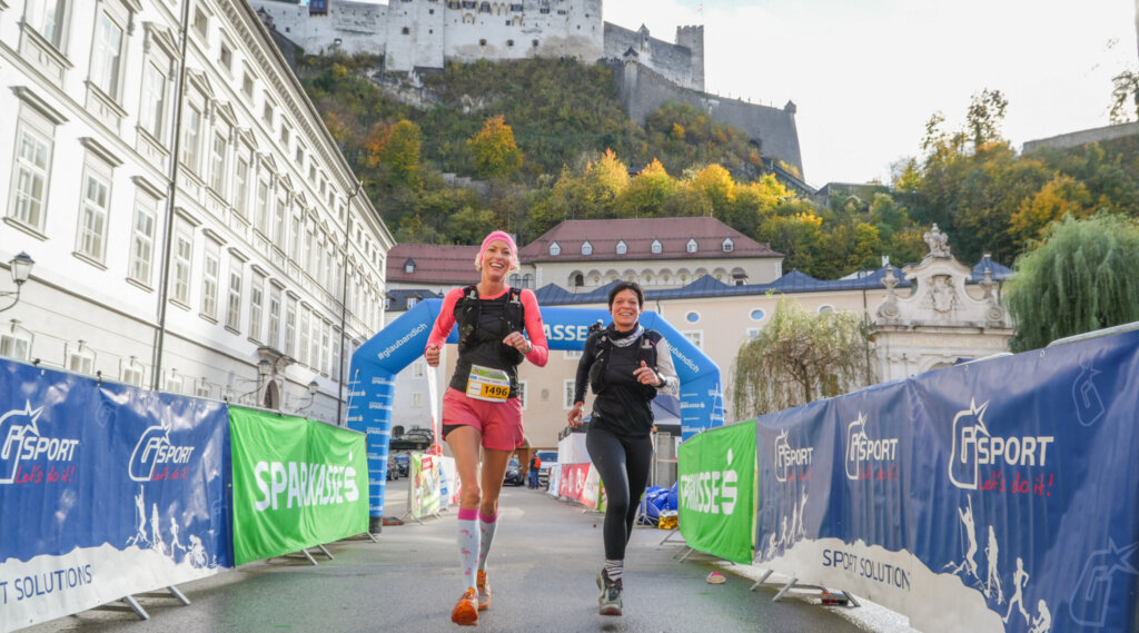 (c) Salzburg Trailrunning Two runners smile as they approach a race finish line, with a castle on a hill in the background. (Enlarged view)