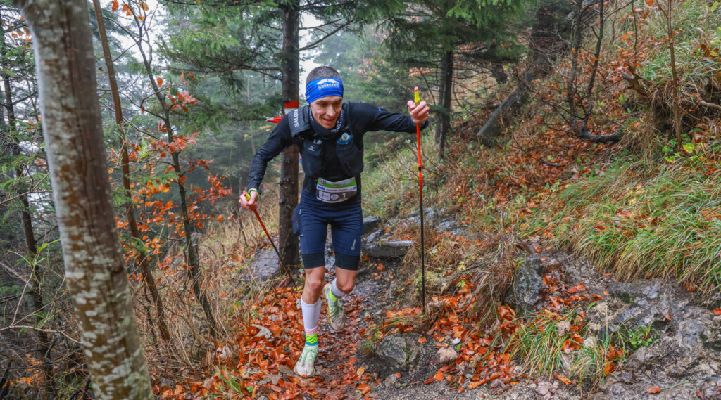 (c) Salzburg Trailrunning A runner with trekking poles climbs a muddy, forested trail during a race on a rainy day. (Enlarged view)