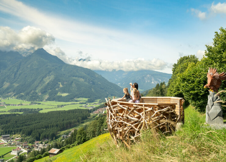Wildkogel Arena Three people admire a scenic mountain valley view from a wooden nest-like lookout platform on a hillside.