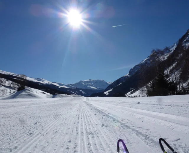 A bright sun shines over a snowy, groomed trail in a mountain valley under a clear blue sky.