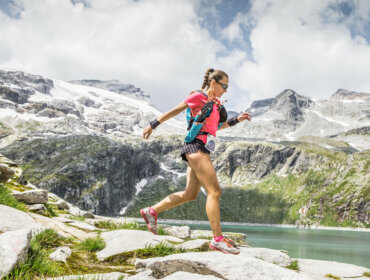 Woman trail running on rocky mountain path near a lake with snow-capped peaks in the background.