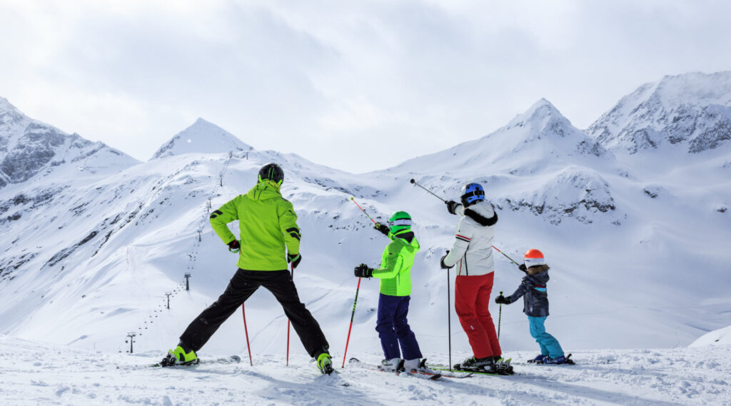 Vier Skifahrer, darunter zwei Kinder, stehen an einem verschneiten Berghang, umgeben von schneebedeckten Gipfeln.