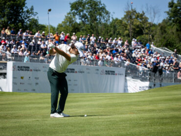 A golfer swings at the ball on a green, with a large crowd watching from stands in the background.