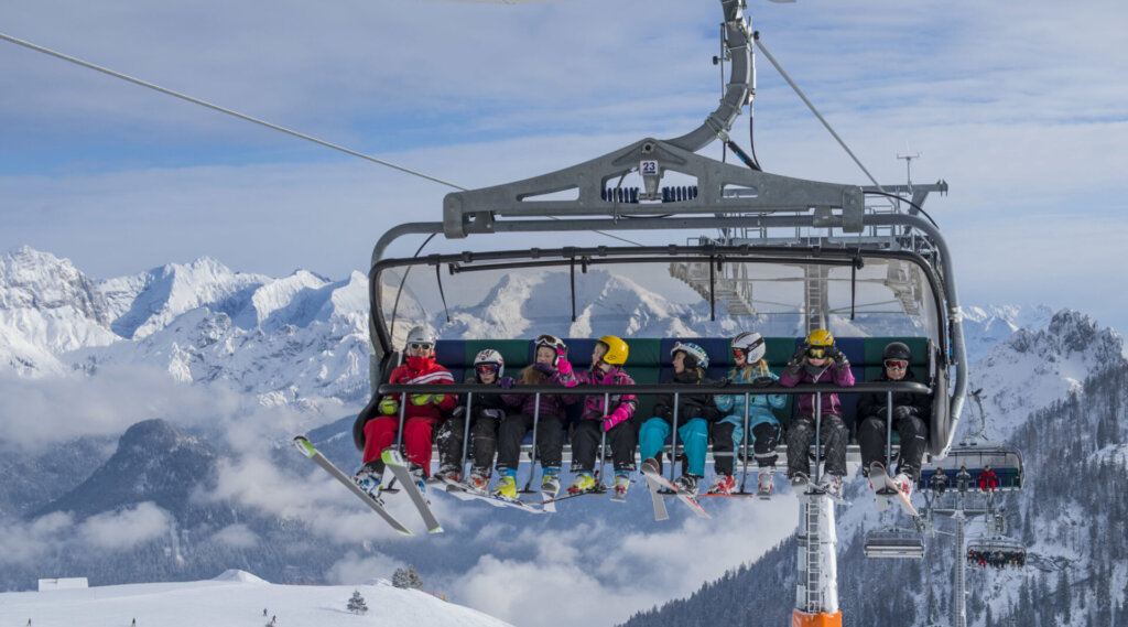 Eine Gruppe von Skifahrern mit Helmen fährt an einem sonnigen Tag mit einem Skilift über verschneite Berge.