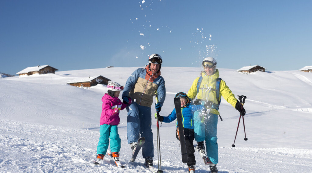 Vierköpfige Familie in bunten Skiklamotten spielt im Schnee auf einem sonnigen, verschneiten Berghang mit Hütten im Hintergrund.
