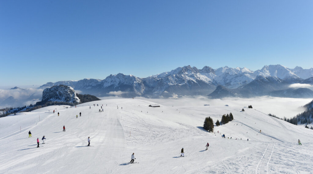 Skifahrer auf einer verschneiten Bergpiste mit klarem Himmel und fernen Berggipfeln im Hintergrund.