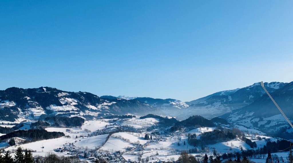Verschneite Skipiste mit Skifahrern, umgeben von Bergen und einem Dorf unter strahlend blauem Himmel.