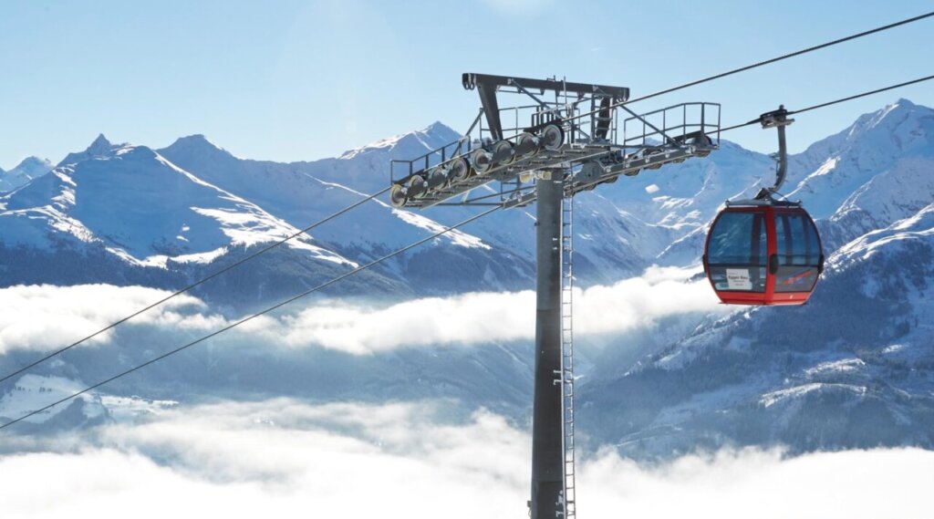 Eine rote Skigondel erhebt sich bei strahlendem Sonnenschein und blauem Himmel über verschneite Berge.