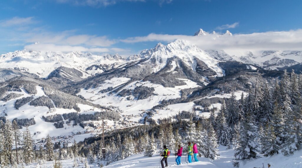 Vier Skifahrer stehen auf einer verschneiten Piste mit Blick auf eine malerische Berglandschaft und ein Dorf unter blauem Himmel.