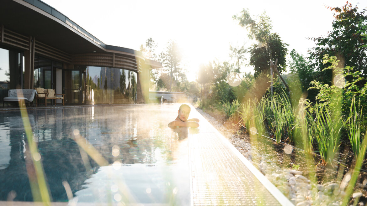 Person relaxing in an outdoor swimming pool beside a modern building on a sunny day, surrounded by greenery. (Enlarged view)