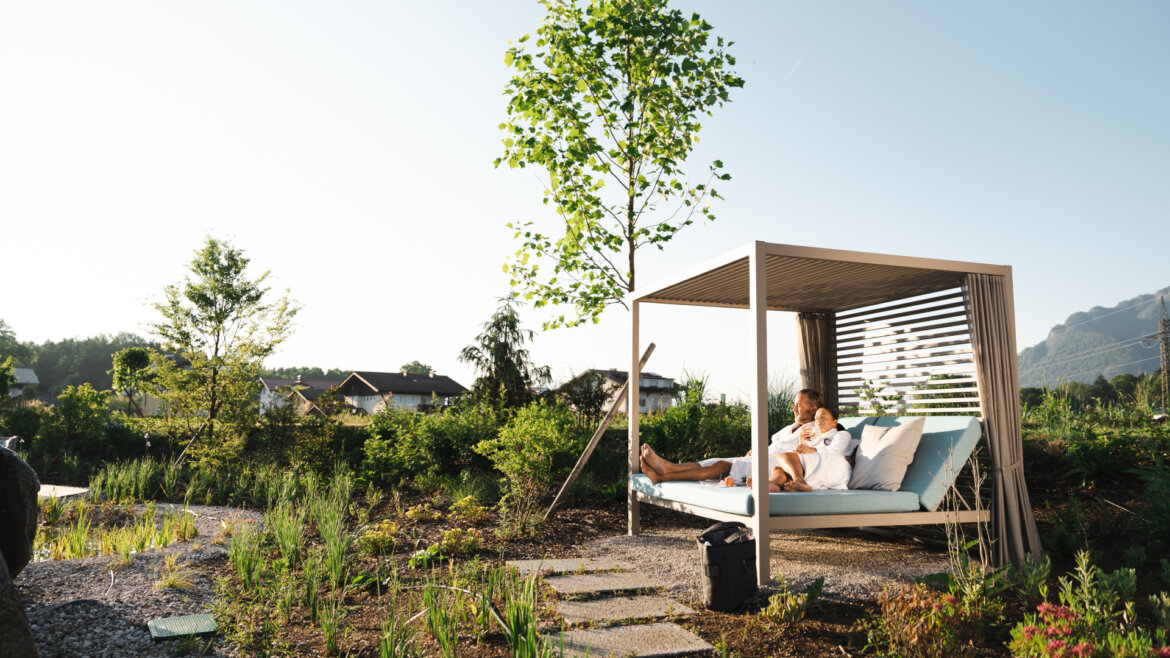 Person relaxing on an outdoor daybed under a canopy, surrounded by greenery and sunlight. (Enlarged view)