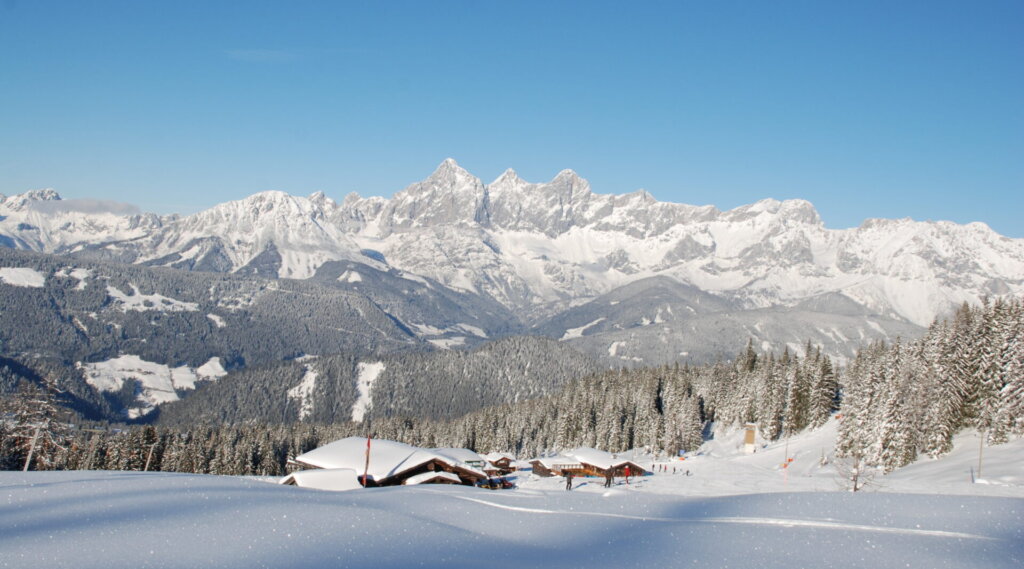 Schneebedeckte Berge und Kiefern unter einem strahlend blauen Himmel, mit Hütten, die in die verschneite Landschaft eingebettet sind.