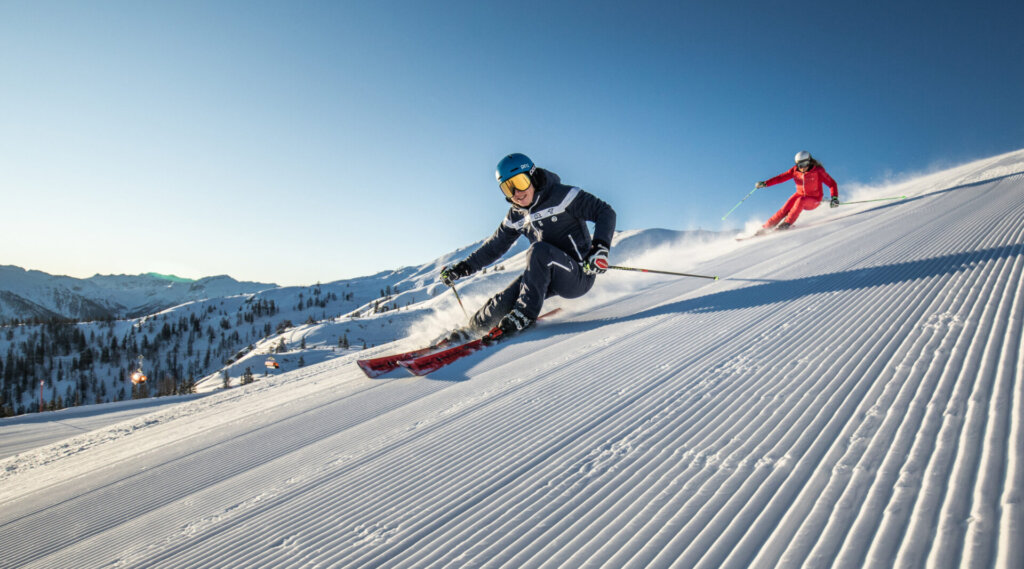 Zwei Skifahrer fahren bei strahlend blauem Himmel in den Bergen eine präparierte Schneepiste hinunter.