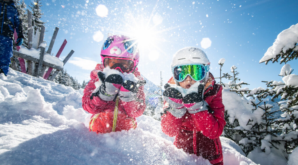 Zwei Kinder in Skikleidung spielen im Schnee, das Sonnenlicht glitzert um sie herum an einem hellen Wintertag.
