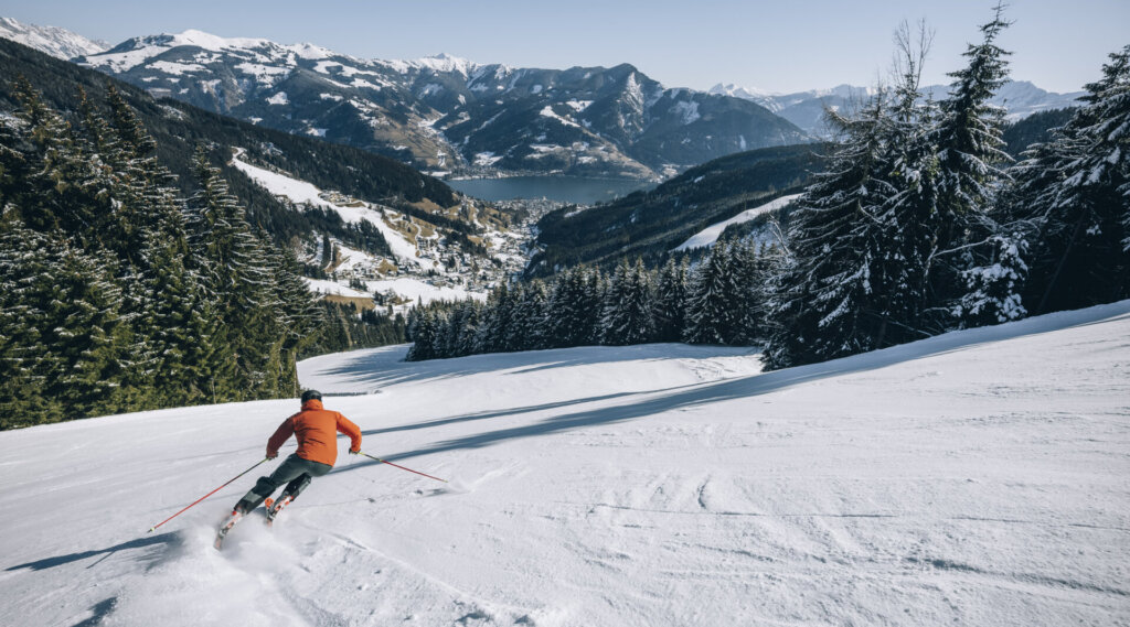 Ein Skifahrer in einer roten Jacke fährt einen verschneiten Berghang hinunter, mit malerischen Bergen und einem See im Hintergrund.