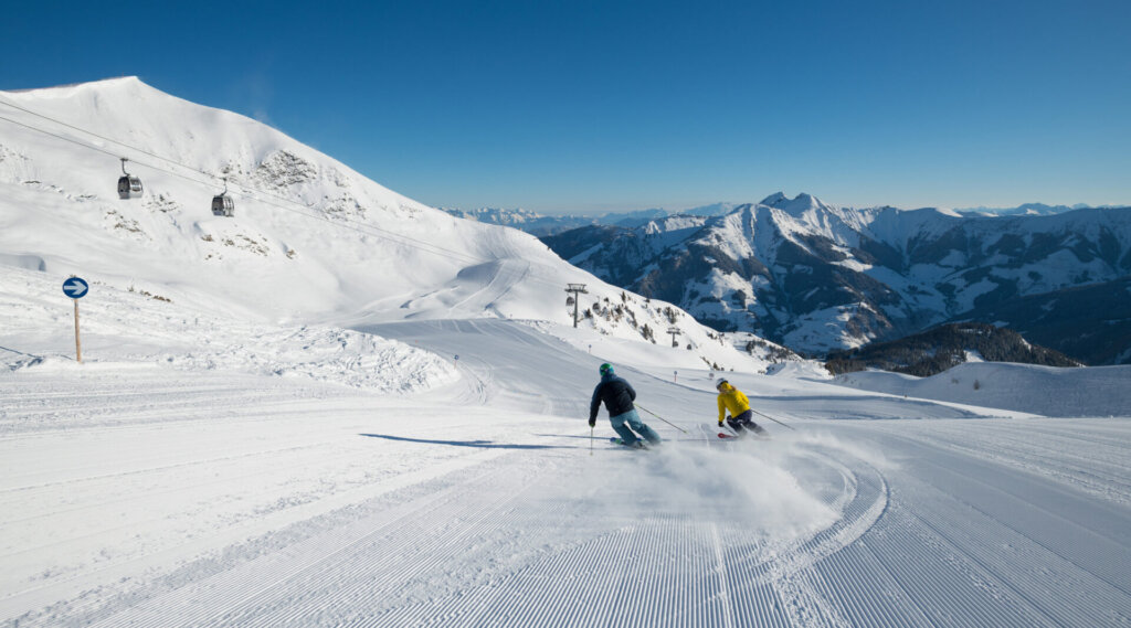 Zwei Skifahrer fahren bei klarem Himmel eine präparierte Schneepiste mit Bergen und Skiliften im Hintergrund hinunter.
