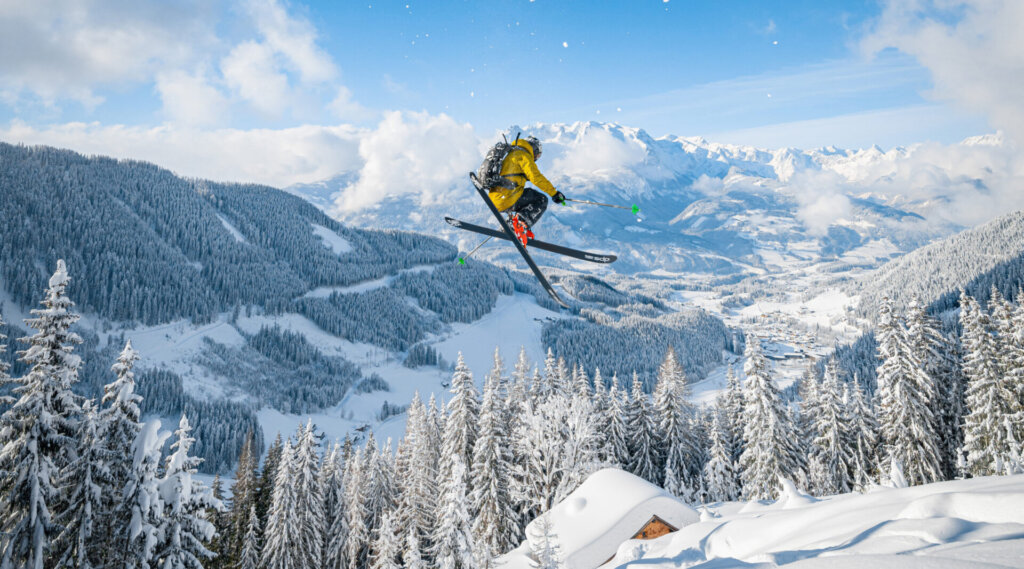 Ein Skifahrer in einer gelben Jacke macht einen Sprungtrick über verschneite Bäume auf einem Berg, mit blauem Himmel und Gipfeln dahinter.